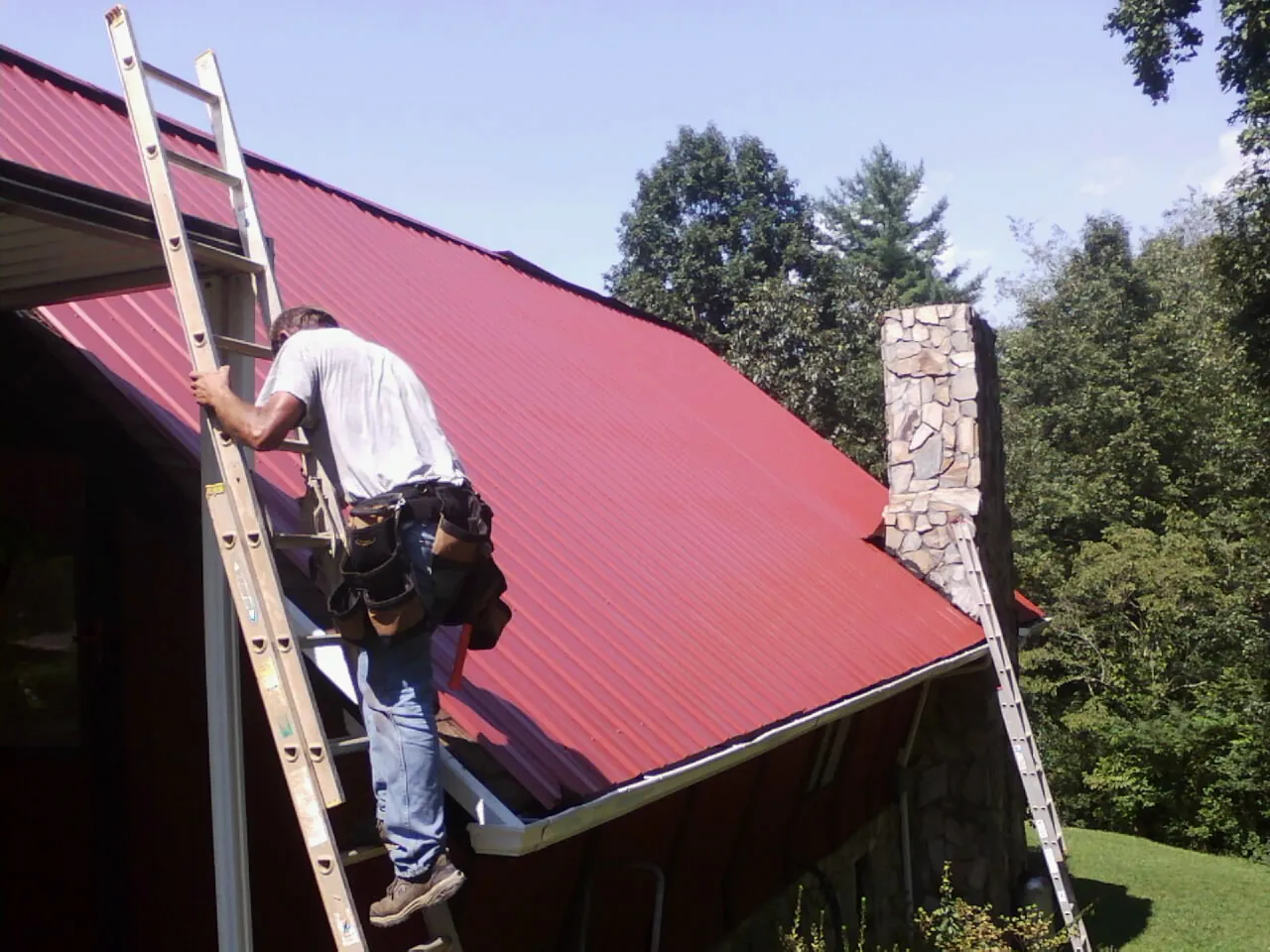 Roofer on a ladder beside a red metal roof and stone chimney — Soffit & Fascia Repair by Roofing Brothers in North Freedom
